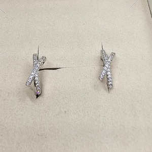 Puede incluir: Un par de pendientes de plata con un diseño de cruz, cada pendiente está incrustado con pequeñas piedras transparentes.