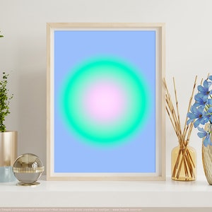 Op de afbeelding: Een ingelijst print met een lichtblauwe achtergrond en een grote, felgroene en roze cirkel in het midden. De print staat op een witte plank met een gouden vaas met blauwe bloemen, een gouden vaas met stokjes en een gouden en blauwe vaas met een plant.