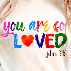 Może przedstawiać: Biały T-shirt z grafiką w kolorach tęczy, na której widnieje napis "you are so loved" z czerwonym sercem w środku słowa "loved". Pod grafiką nadrukowany jest tekst "john 3:16".
