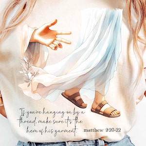 May include: Cream-colored t-shirt featuring a watercolor illustration of a figure in flowing white robes and sandals. The image includes a hand gesture and text that reads, "If you're hanging on by a thread, make sure it's the hem of his garment. matthew 9:20-22."
