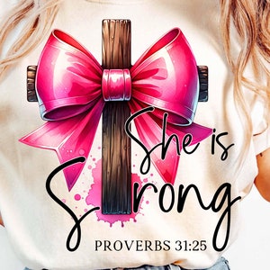 May include: A white t-shirt with a pink bow tied around a brown wooden cross. The text "She is Strong" and "Proverbs 31:25" are printed below the cross.