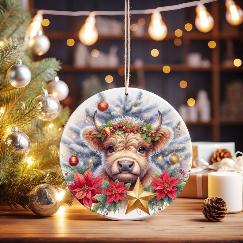 May include: Round Christmas ornament featuring a Highland cow with a festive wreath, surrounded by poinsettias, a gold star, and Christmas tree branches. The ornament hangs from a string, with a Christmas tree and lights in the background.
