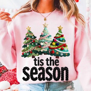 May include: A pale pink sweatshirt with three embroidered Christmas trees, each topped with a gold star. The trees are adorned with festive decorations. The words "tis the season" are embroidered in black below the trees.