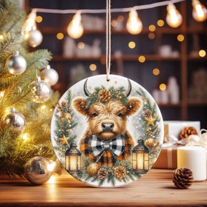 May include: A round Christmas ornament featuring a Highland cow with a plaid bow tie and a wreath of pine needles and pine cones. The ornament is hanging in front of a Christmas tree with silver ornaments and lights.