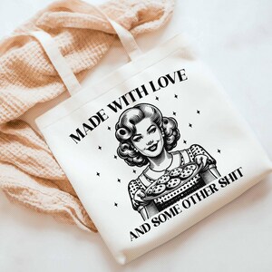 Pode incluir: Saco tote branco com o texto "MADE WITH LOVE AND SOME OTHER SHIT" e uma ilustra&ccedil;&atilde;o em estilo vintage de uma mulher segurando uma bandeja de biscoitos. O saco tem al&ccedil;as creme e est&aacute; sobre um fundo neutro.