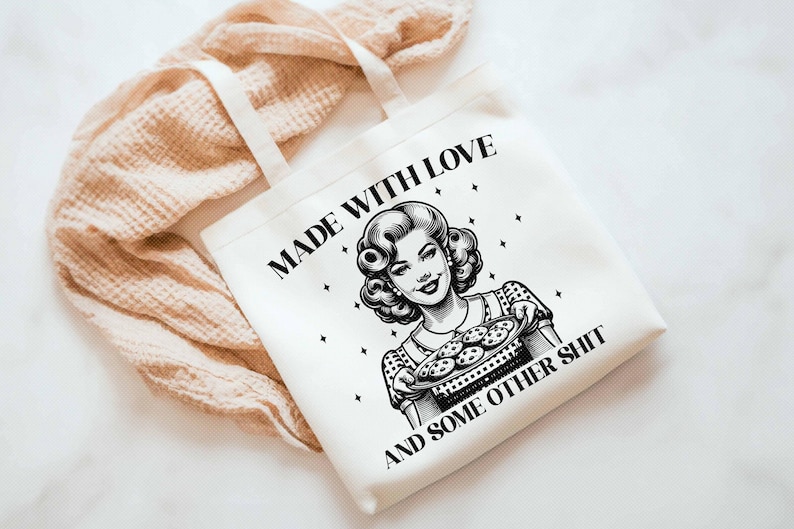 Pode incluir: Saco tote branco com o texto "MADE WITH LOVE AND SOME OTHER SHIT" e uma ilustra&ccedil;&atilde;o em estilo vintage de uma mulher segurando uma bandeja de biscoitos. O saco tem al&ccedil;as creme e est&aacute; sobre um fundo neutro.