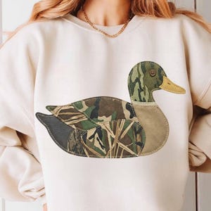 Peut inclure: Sweat-shirt crème avec une grande application de canard. Le motif du canard intègre des motifs de camouflage verts, bruns et beiges. Le bec du canard est jaune, et le design est cousu sur le sweat-shirt.