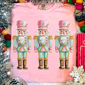 以下が含まれることがあります： 3体のカラフルなくるみ割り人形のイラストが描かれたピンク色のTシャツ。各くるみ割り人形はピンクの帽子、青いコート、茶色のブーツを着用しています。Tシャツのタグには「Comfort Colors」の文字があります。