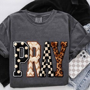 Op de afbeelding: Donkergrijze sweatshirt met het woord "PRAY" in een patchwork-ontwerp. De letter "P" is zwart met witte stippen, "R" is cr&egrave;me met een bruin zigzagpatroon, "A" is zwart-wit geblokt en "Y" is luipaardprint.