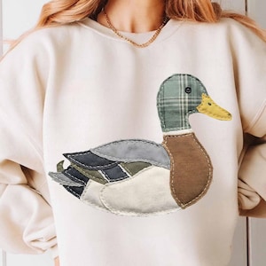 Peut inclure: Sweat-shirt crème avec un motif de canard en patchwork. Le canard a une tête à carreaux verts, un bec jaune et des ailes faites de différentes nuances de bleu, gris et blanc. Le corps est marron et le motif est cousu sur le sweat-shirt.
