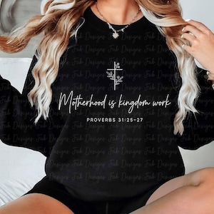May include: Black sweatshirt with the text "Motherhood is kingdom work" and a cross design with floral accents. The text "PROVERBS 31:25-27" is below the main text. The sweatshirt is a crew neck style.