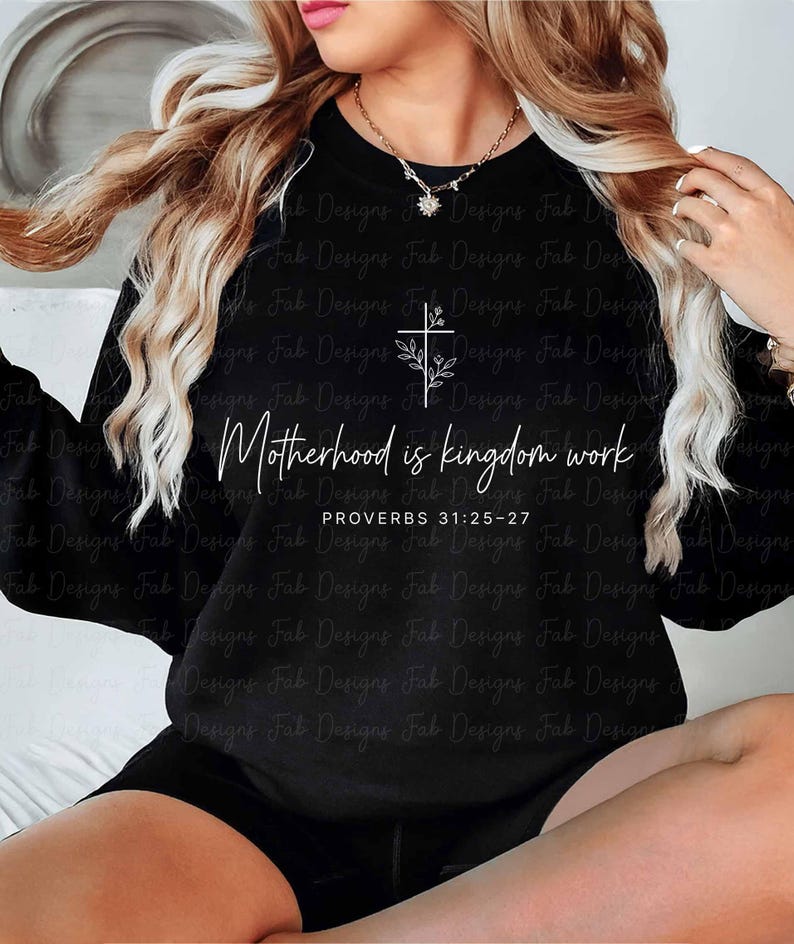 May include: Black sweatshirt with the text "Motherhood is kingdom work" and a cross design with floral accents. The text "PROVERBS 31:25-27" is below the main text. The sweatshirt is a crew neck style.