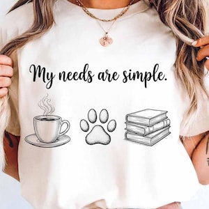 May include: Cream-colored t-shirt with the text "My needs are simple." Below the text are illustrations of a coffee cup, a paw print, and a stack of books. The design is in black and white.