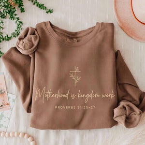 May include: A brown sweatshirt with the text "Motherhood is kingdom work" and "Proverbs 31:25-27" in a cursive font. A cross with floral accents is above the text. The sleeves are rolled up.