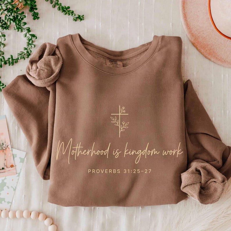 May include: A brown sweatshirt with the text "Motherhood is kingdom work" and "Proverbs 31:25-27" in a cursive font. A cross with floral accents is above the text. The sleeves are rolled up.