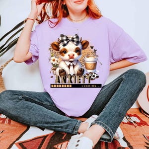May include: A light purple t-shirt featuring a cartoon cow with a bow tie, holding a coffee cup. The text "ANXIETY LOADING..." is below the cow. The cow is surrounded by flowers and a leopard print background.