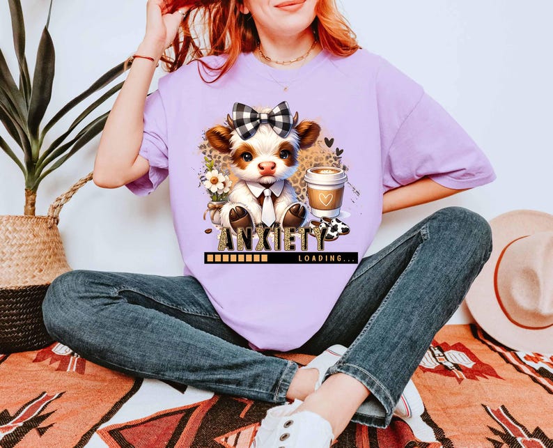 May include: A light purple t-shirt featuring a cartoon cow with a bow tie, holding a coffee cup. The text "ANXIETY LOADING..." is below the cow. The cow is surrounded by flowers and a leopard print background.