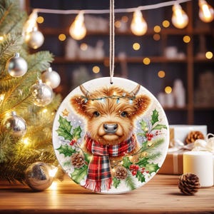 May include: A round Christmas ornament featuring a Highland cow with a red and black plaid scarf, adorned with holly, berries, and pinecones. The cow has a string of lights around its horns. The ornament hangs from a string.