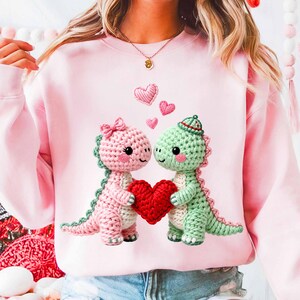 May include: A light pink sweatshirt featuring two crocheted dinosaur figures holding a red heart. One dinosaur is pink with a bow, the other is green with a hat. Small pink hearts float above them.
