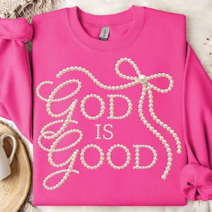 May include: A vibrant pink sweatshirt featuring the words "God is Good" crafted from white faux pearls. The design incorporates a pearl bow and pearl strands. The sweatshirt has long sleeves.
