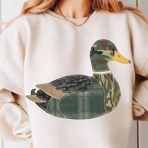Peut inclure: Sweat-shirt crème avec une grande application de canard. Le motif du canard intègre diverses textures et motifs, notamment le camouflage, le tartan et des couleurs unies. Le canard a un bec jaune et est cousu sur le sweat-shirt.