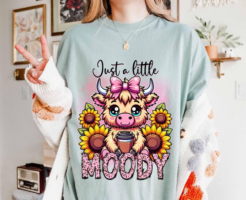 May include: A light blue sweatshirt with a cartoon highland cow wearing a pink bow and holding a cup of coffee. The text "Just a little MOODY" is printed on the shirt.