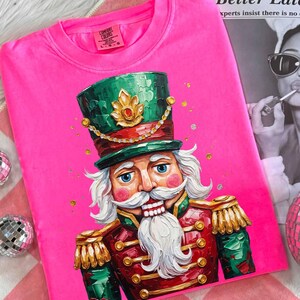 Może przedstawiać: Jasnoróżowa bluza Comfort Colors z szczegółową ilustracją żołnierza Dziadka do Orzechów. Dziadek do Orzechów ma zielony kapelusz, czerwono-złoty mundur i białe wąsy. Widoczne są złote akcenty i logo Comfort Colors.