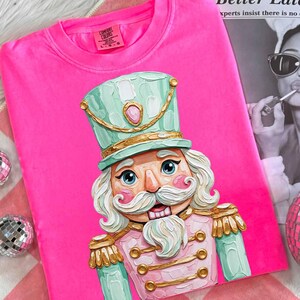 May include: A bright pink Comfort Colors t-shirt featuring a detailed illustration of a nutcracker soldier. The nutcracker wears a mint green hat and jacket with gold accents. The shirt is laid on a pink and white checkered surface.