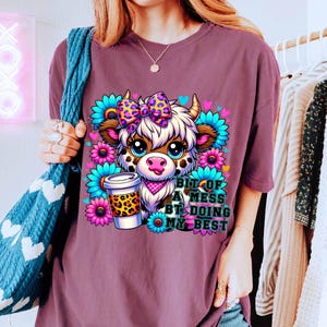 May include: Purple t-shirt with a cartoon cow wearing a bow and holding a coffee cup. The text on the shirt reads "Bit of a mess but doing my best".