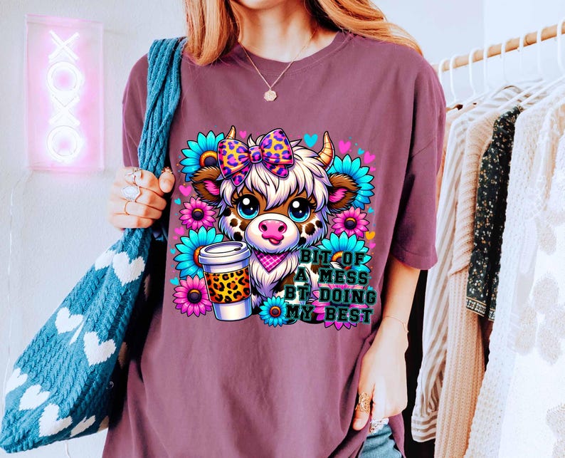 May include: Purple t-shirt with a cartoon cow wearing a bow and holding a coffee cup. The text on the shirt reads "Bit of a mess but doing my best".