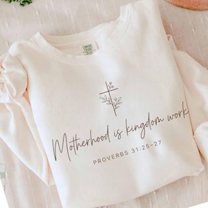 May include: Cream-colored sweatshirt with the text "Motherhood is kingdom work" and "PROVERBS 31:25-27" in a brown script. A cross with floral accents is above the text. The sweatshirt has a crew neck and long sleeves.