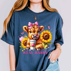 May include: Navy blue t-shirt featuring a cartoon calf with a pink bow, drinking from a coffee cup, and surrounded by sunflowers and butterflies. The text "STAY SASSY" is printed in pink below the calf.