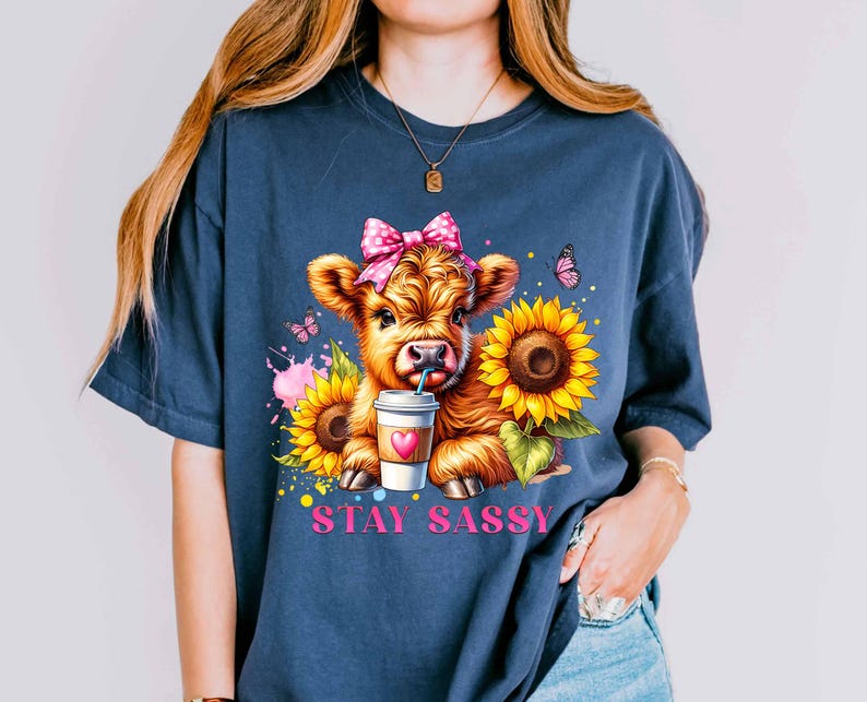 May include: Navy blue t-shirt featuring a cartoon calf with a pink bow, drinking from a coffee cup, and surrounded by sunflowers and butterflies. The text "STAY SASSY" is printed in pink below the calf.