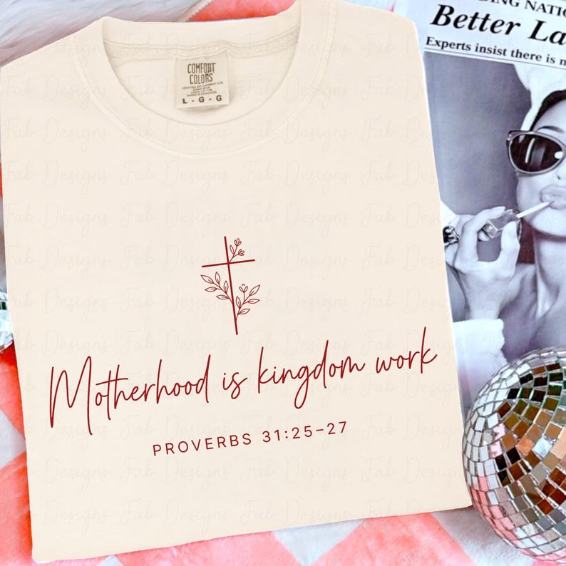 May include: Cream-colored t-shirt with the words "Motherhood is kingdom work" and the Bible verse Proverbs 31:25-27 in a burgundy script. A burgundy cross and floral design is above the text. The shirt has a Comfort Colors tag.