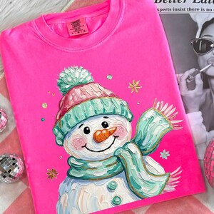 May include: A bright pink Comfort Colors t-shirt featuring a smiling snowman illustration. The snowman wears a teal and pink hat and scarf, with a carrot nose and gold accents. The shirt is a size large.