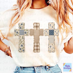 May include: A cream-colored t-shirt featuring a graphic of three crosses arranged side-by-side. The left cross has a blue floral and gingham pattern, the center cross has a tan plaid and striped pattern, and the right cross has a blue polka dot and striped pattern with small cross icons.