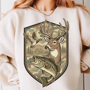 Puede incluir: Sudadera color crema con un gráfico temático de caza. El diseño incluye un ciervo, un pato en vuelo y un pez, sobre un fondo de camuflaje dentro de una forma de escudo. El gráfico tiene tonos marrones, verdes y beige.
