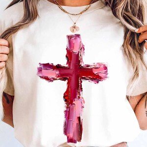 May include: Cream-colored t-shirt featuring a large, textured cross design in shades of red and pink. The cross appears to be painted with visible brushstrokes, giving it an artistic, expressive look. The shirt is a casual wear item.