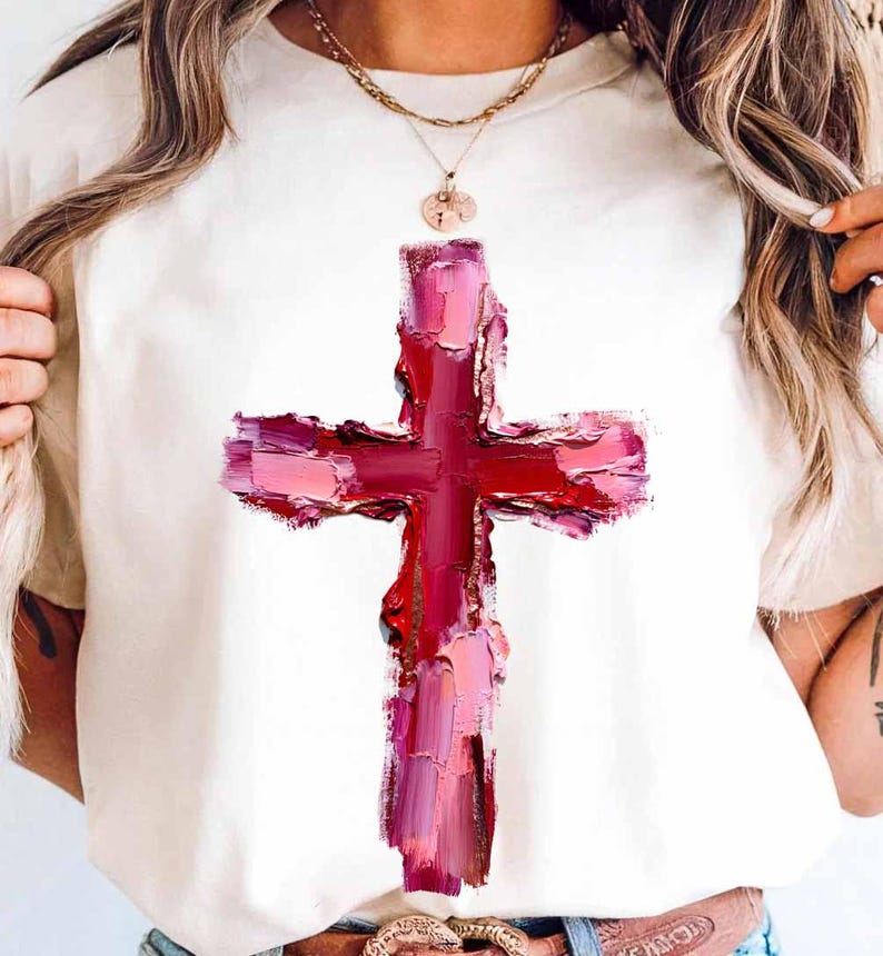May include: Cream-colored t-shirt featuring a large, textured cross design in shades of red and pink. The cross appears to be painted with visible brushstrokes, giving it an artistic, expressive look. The shirt is a casual wear item.
