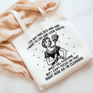 K&ouml;nnte beinhalten: Wei&szlig;er Canvas-Tote-Bag mit schwarzem Text und einer Illustration im Vintage-Stil einer Frau mit einem Besen. Der Text lautet: "I'VE GOT PMS OCD AND ADD I WANT TO CRY AND LOOK PRETTY...RIGHT NOW AS I'M CLEANING."