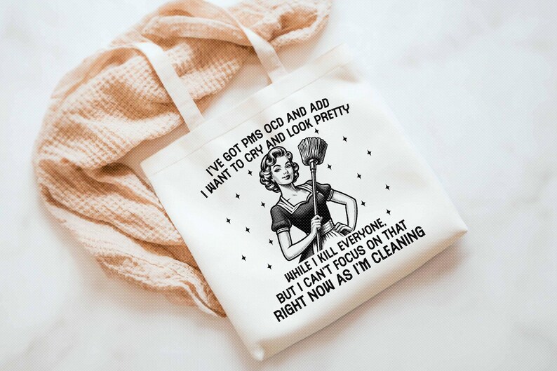 K&ouml;nnte beinhalten: Wei&szlig;er Canvas-Tote-Bag mit schwarzem Text und einer Illustration im Vintage-Stil einer Frau mit einem Besen. Der Text lautet: "I'VE GOT PMS OCD AND ADD I WANT TO CRY AND LOOK PRETTY...RIGHT NOW AS I'M CLEANING."