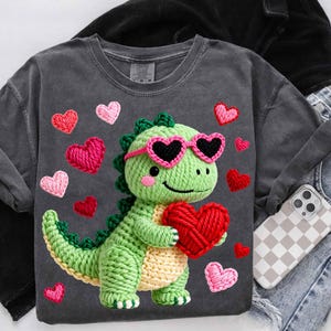 May include: Dark gray t-shirt featuring a crocheted green dinosaur wearing pink heart-shaped sunglasses and holding a red heart. Pink and red crocheted hearts are scattered around the dinosaur. The shirt is paired with denim jeans and a phone case.