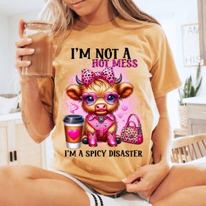 May include: A tan t-shirt with a cartoon cow wearing pink boots, sunglasses, and a pink bow. The text on the shirt reads "I'm not a hot mess, I'm a spicy disaster."