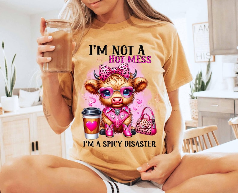 May include: A tan t-shirt with a cartoon cow wearing pink boots, sunglasses, and a pink bow. The text on the shirt reads "I'm not a hot mess, I'm a spicy disaster."