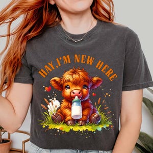 May include: Dark gray t-shirt featuring a cartoon calf with orange fur, holding a bottle. The text "HAY, I'M NEW HERE" is arched above the calf. The calf is sitting in grass with hearts and flowers.