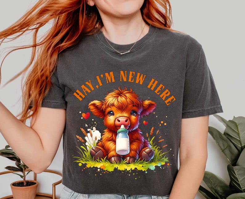May include: Dark gray t-shirt featuring a cartoon calf with orange fur, holding a bottle. The text "HAY, I'M NEW HERE" is arched above the calf. The calf is sitting in grass with hearts and flowers.