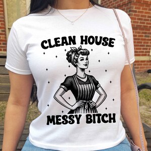 K&ouml;nnte beinhalten: Wei&szlig;es T-Shirt mit dem schwarzen Text "CLEAN HOUSE MESSY BITCH", das eine Vintage-Illustration einer Frau mit Sch&uuml;rze zeigt. Das Design enth&auml;lt Sternakzente um die Illustration. Das Shirt ist aus weichem Material.