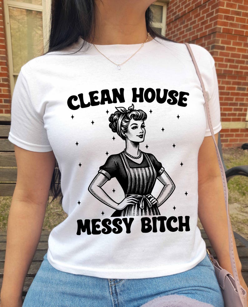 K&ouml;nnte beinhalten: Wei&szlig;es T-Shirt mit dem schwarzen Text "CLEAN HOUSE MESSY BITCH", das eine Vintage-Illustration einer Frau mit Sch&uuml;rze zeigt. Das Design enth&auml;lt Sternakzente um die Illustration. Das Shirt ist aus weichem Material.