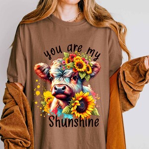 May include: Brown t-shirt featuring a colorful illustration of a cow adorned with sunflowers and a floral crown. The text "you are my Shunshine" is displayed below the cow. The shirt has short sleeves.