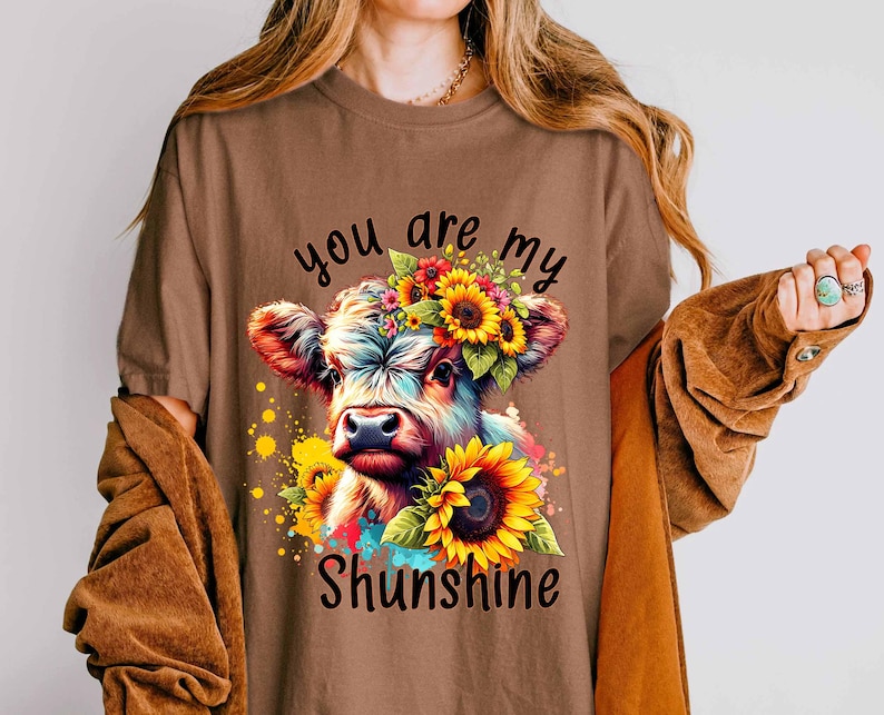 May include: Brown t-shirt featuring a colorful illustration of a cow adorned with sunflowers and a floral crown. The text "you are my Shunshine" is displayed below the cow. The shirt has short sleeves.